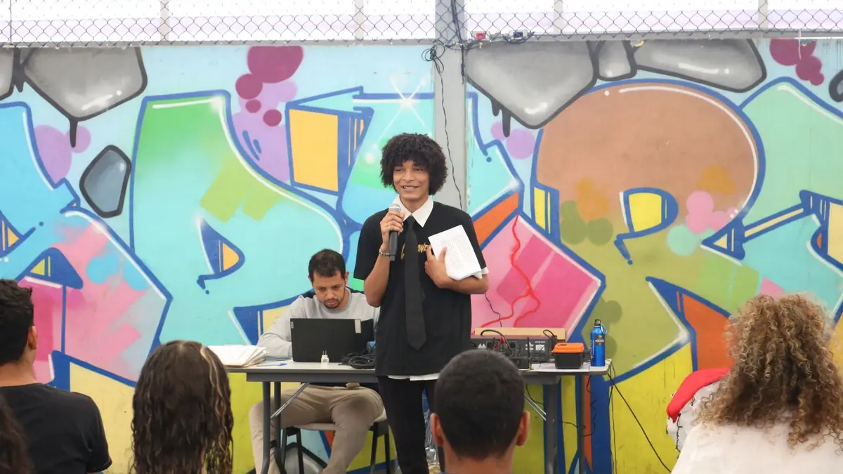 Jovem da Despertar falando ao microfone diante de um painel de grafite